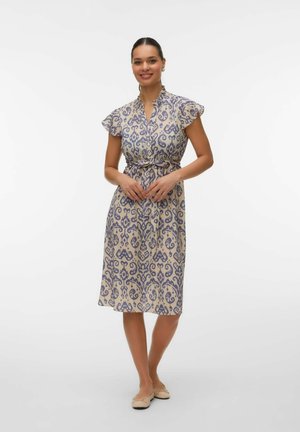 Uśmiechnięta kobieta stojąca z złączonymi rękami, ubrana w beżową sukienkę do kolan z fioletowymi abstrakcyjnymi wzorami oraz beżowe baleriny na białym tle.