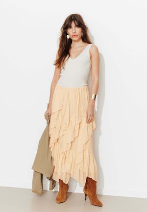 Femme aux longs cheveux bruns portant un haut blanc sans manches, une jupe à volants couleur pêche, des bottines marron à talons, tenant une veste beige, debout contre un mur blanc.