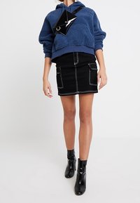 Sudadera de forro polar azul con una falda corta de mezclilla negra con bolsillos frontales, combinada con botas de tobillo negras y un bolso geométrico negro.