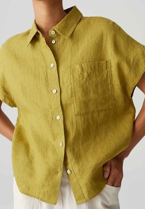 Persoon die een loszittend olijfgroen linnen shirt met korte mouwen draagt, voorzien van een borstzak, witte knopen en witte broek, handen in de zakken.