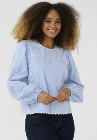 Blusa azzurra chiara con maniche a sbuffo, orlo ondulato e cuciture decorative bianche. Realizzata in tessuto morbido con polsini arricciati.