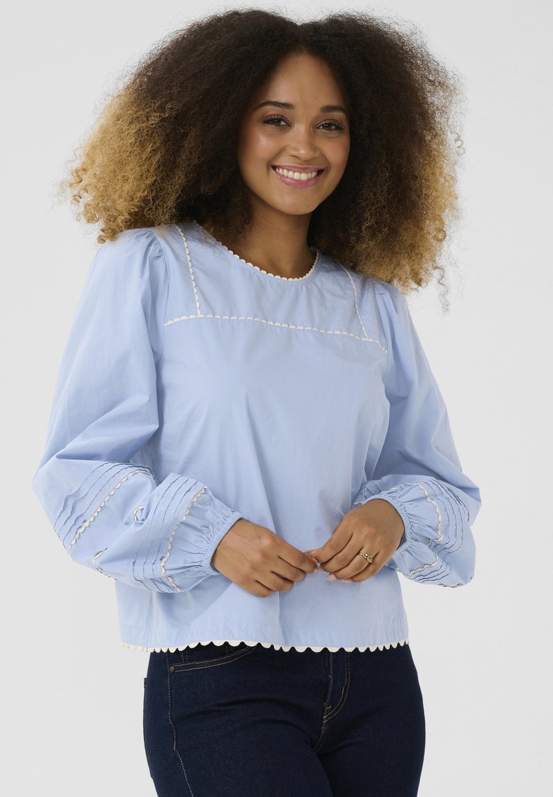 Blusa azzurra chiara con maniche a sbuffo, orlo ondulato e cuciture decorative bianche. Realizzata in tessuto morbido con polsini arricciati.