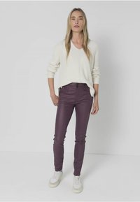 Crème V-hals trui, ribbelstructuur, gecombineerd met aansluitende, donkerpaarse broek. Model staat in witte sneakers voor een effen witte achtergrond.