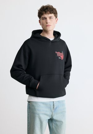 Tânăr purtând un hanorac negru cu grafică de trandafir roșu și blugi albaștri deschiși, stând cu mâinile în buzunarul hanoracului, pe fundal simplu.