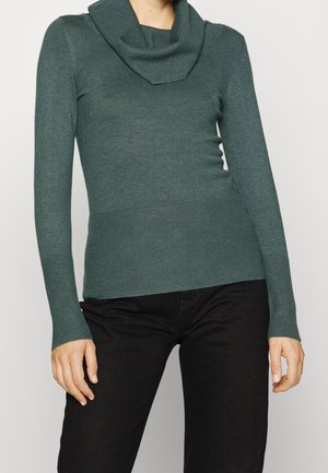 Vrouw draagt een tealkleurige trui met lange mouwen, een col en geribbelde manchetten, gecombineerd met een zwarte broek, staand tegen een lichte achtergrond.