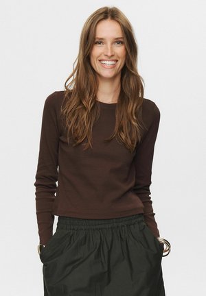 Brown long-sleeve top with a round neckline, fitted design. Paired with a black, elastic-waist, gathered skirt. Model smiles, hands in pockets.