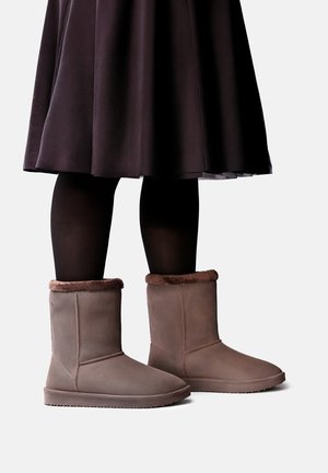Jambes portant des collants noirs, une jupe sombre et des bottes en daim marron mi-mollet avec une doublure en fourrure, sur un fond clair uni.