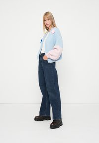 Ανοιχτό μπλε πλεκτό cardigan με ροζ φουσκωτά μανίκια και λουλούδιες εφαρμογές, συνδυασμένο με σκούρα μπλε φαρδιά τζιν παντελόνια και χοντρά μαύρα παπούτσια.