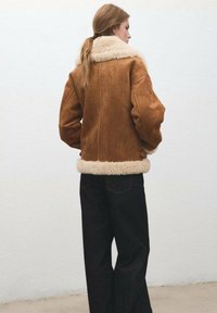 Bruine suède jas met een zachte, crèmekleurige kraag en zoom. Heeft een ontspannen pasvorm, lange mouwen en een stoftextuur die contrasteert met gladheid.