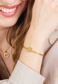 Gouden armband met een ronde bedel met uitgesneden ontwerp. Draag op een lichtgekleurde pols, aangevuld met een lichte, textuur blouse.