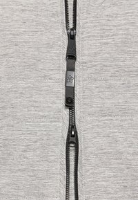 Light grey zip-up jacket made of textured fabric with a central black zipper featuring branded pull tab and stitching accents.