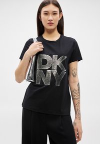Jovem mulher de cabelos compridos e escuros, vestindo camisa preta da DKNY e calças pretas, segurando uma mala prateada metálica, com tatuagens detalhadas no braço direito.