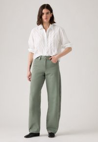Une femme se tient avec une main dans la poche, portant une blouse blanche à manches longues et un pantalon ample vert sauge avec des chaussures noires.