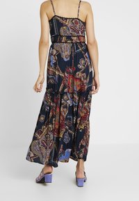 Kvinna i en ärmlös, marinblå maxiklänning med flerfärgade paisleymönster och lila klacksandaler, stående mot en vit bakgrund.