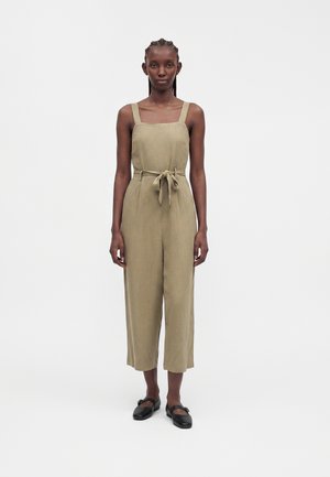 Kvinde stående vendt mod fronten iført en ærmeløs beige buksedragt med taljebånd og sorte flade sko mod en ensfarvet baggrund.