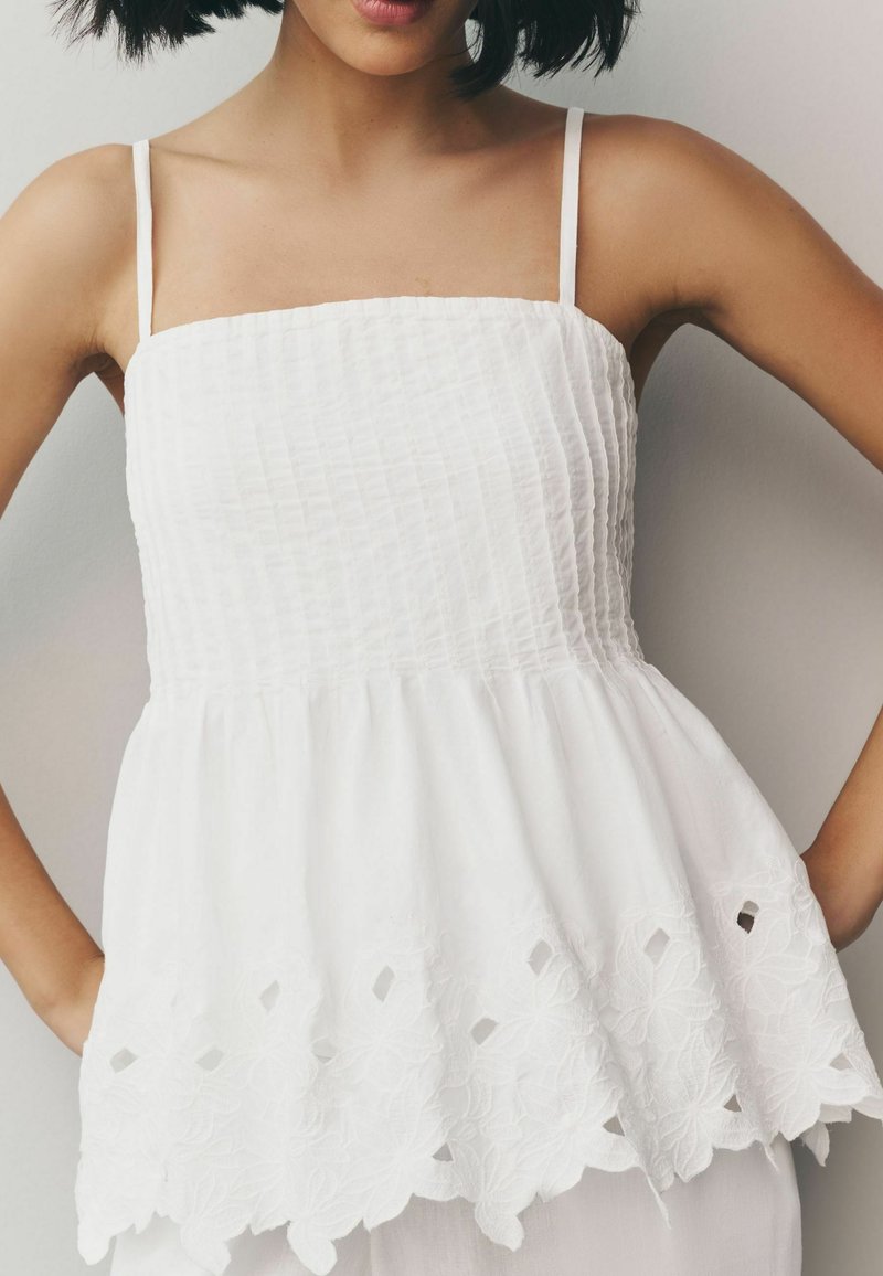 Haut peplum blanc à fines bretelles, panel supérieur texturé et détail de bordure florale brodé. Tissu doux avec un léger éclat.