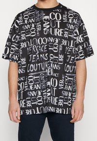 Černé tričko s krátkým rukávem s grafickým potiskem slov "Versace Jeans Couture" v bílé barvě, zobrazující různé styly písma a orientace.
