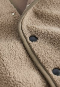 Beige texturerat tyg med en fleece-liknande framtoning, med en slät olivgrön kant och svarta tryckknappar.