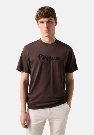 Junger Mann trägt ein dunkelbraunes T-Shirt mit besticktem Text und hellbeigen Hosen, steht vor einem schlichten hellen Hintergrund.