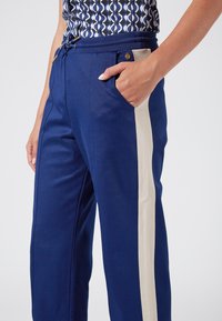 Marina byxor med en kontrasterande krämfärgad sidstripe, dragsko i midjan och sidoficka; tillverkade av ett smidigt, lättviktsmaterial.