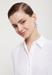Hvid skjorte med krave, der har en glat tekstur. Modellens ørepynt består af store sølvfarvede cirkulære øreringe med en struktureret overflade.