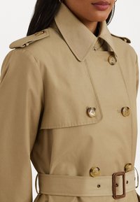 Camel-colored trench coat featuring a double-breasted design, large buttons, a wide collar, and a matching belt at the waist.