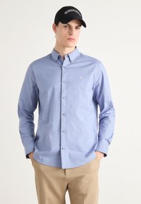 Camisa azul clara de botões com colarinho, mangas longas e um pequeno logotipo dourado; combinada com calças bege e um boné preto com letras brancas.