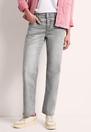 Vrouw draagt lichtgrijze high-waist jeans met dubbele knoopsluiting, witte sneakers, wit topje en roze jas, staat met één hand in de zak.