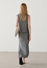 Frau mit langen blonden Haaren, die weggeht, in einem ärmellosen grauen Strickkleid, schwarzen Schieberandsandalen und mit einer beigefarbenen Umhängetasche.