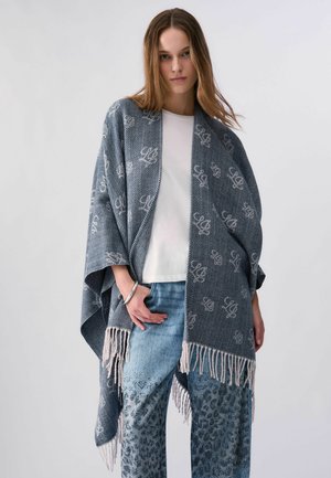 Jonge vrouw die een grijs patroonrijk omslagdoek met franjes draagt over een wit overhemd en blauwe patroonrijke spijkerbroek, staand tegen een effen achtergrond.