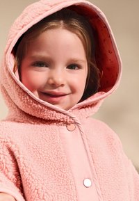 Różowy płaszcz z polaru z dużym kapturem, posiadający gładką podszewkę wewnętrzną i dwa zatrzaski. Materiał ma miękką, teksturowaną powierzchnię.