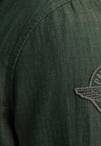 Chemise vert foncé avec un motif en chevrons, ornée de détails en patch brodé gris et de coutures d'épaule structurées.