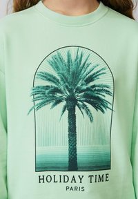 Miętowa bluza z dużą czarną grafiką palmy w łuku, z napisami "HOLIDAY TIME PARIS" poniżej.