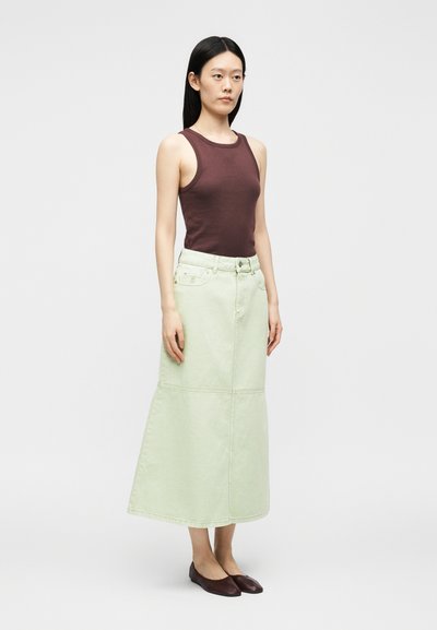 Femme portant un haut marron sans manches, une longue jupe verte claire taille haute et des ballerines marron foncé, debout devant un fond clair uni.