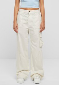 Beige cargo bukser med løs pasform, med sidelommer, subtile syningsdetaljer og lynlås ved manchetterne, parret med hvide sneakers.