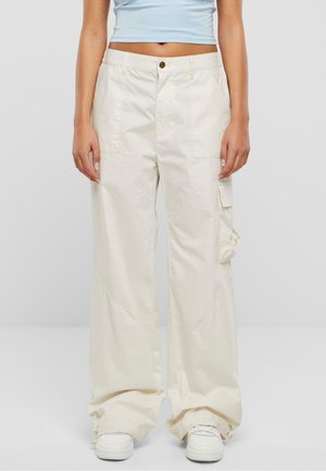 Beige cargo bukser med løs pasform, med sidelommer, subtile syningsdetaljer og lynlås ved manchetterne, parret med hvide sneakers.