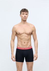 Mustat boxerit punaisella vyötäröllä, jossa on "TOMMY HILFIGER" -logo. Kangas on joustavaa ja sileää, ilman näkyviä kuvioita tai koristeita.