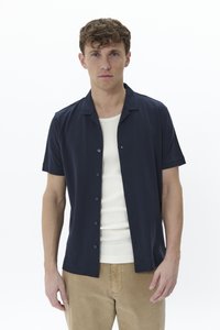 Chemise à manches courtes, avec des boutons, en tissu bleu marine au toucher doux, dotée d'un col évasé et d'une coupe ajustée, portée par-dessus un débardeur blanc à côtes.