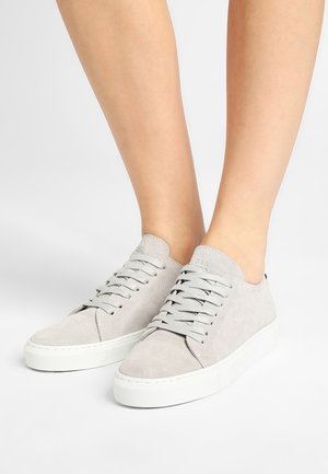 Stopy w lekkich jasnoszarych niskich trampkach z zamszu z białymi gumowymi podeszwami i dopasowanymi szarymi sznurowadłami na białym tle.