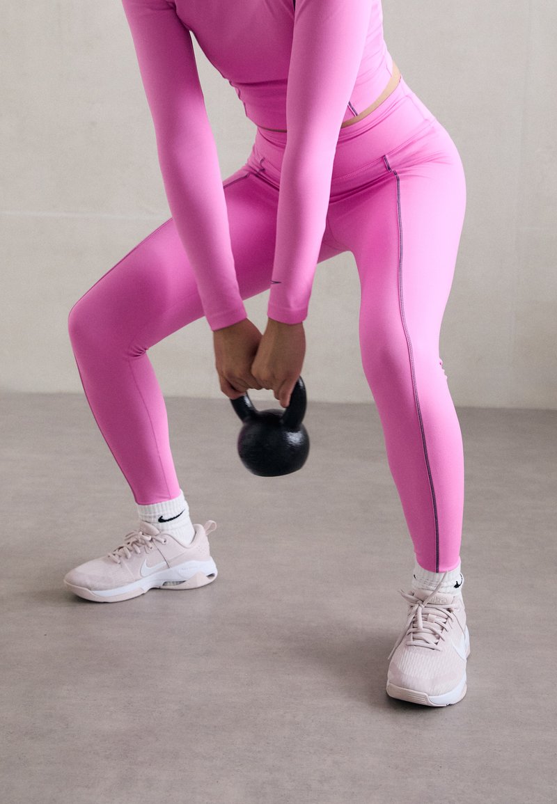Ružový dlhý rukáv top a zodpovedajúce legíny, oboje tesne priliehajúce, s čiernymi akcentmi. Drží čierny kettlebell a má na sebe svetlo ružové športové topánky.