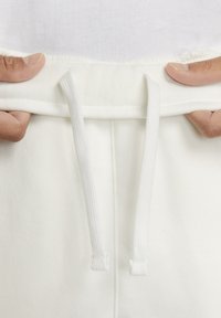 White sweatpants featuring a drawstring waistband, flat stitching, and a smooth cotton texture. Hands are holding the waistband.