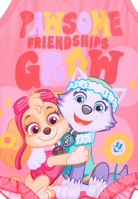 Le maillot de bain rose présente des chiens de dessin animé, l'un avec des lunettes de natation et l'autre en tenue bleue, avec le texte "DES AMITIÉS INCROYABLES GRANDISSENT" dans des couleurs vives.