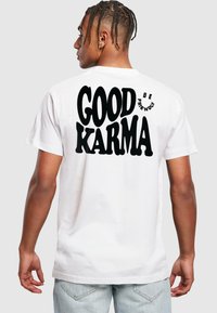 Bílá bavlněná tričko s velkým černým textem na zadní straně, který říká "DOBRÁ KARMA" s nápisem "BUĎ PŘÁTELSKÝ" pod ním menším zakřiveným písmem.