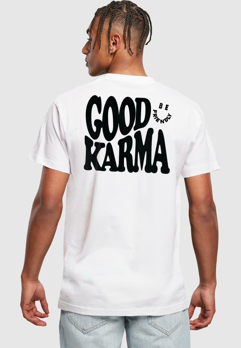 Bílá bavlněná tričko s velkým černým textem na zadní straně, který říká "DOBRÁ KARMA" s nápisem "BUĎ PŘÁTELSKÝ" pod ním menším zakřiveným písmem.
