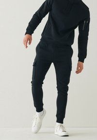 Uomo che indossa pantaloni jogger neri con tasche laterali, felpa nera, calzini bianchi e scarpe da ginnastica bianche, in piedi contro un muro bianco semplice.