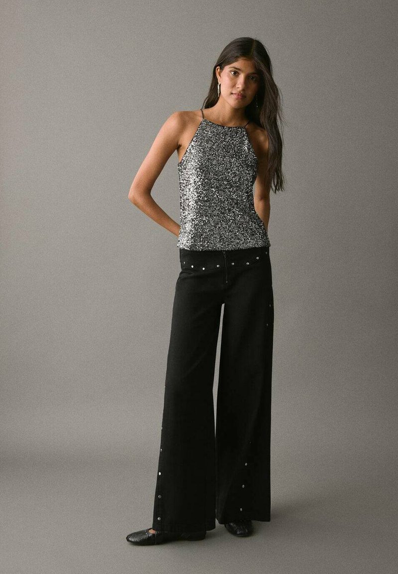 Strieborný flitrový top s halter strihom v kombinácii s vysokými širokými čiernymi nohavicami, ktoré majú zlato vrecká po stranách a hladkú textúru.