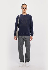Marine sweatshirt med slidte print, grå jeans, hvide sneakers og en lys grå crossover taske. Sorte solbriller tilføjer et accent.