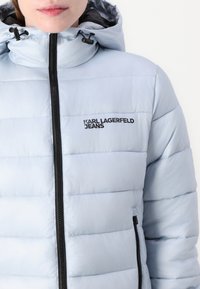 Lyseblå puffjakke med glidelåsåpning, justerbar hette, sidelommer og sort logo-detaljering. Glatt, quiltet tekstur.