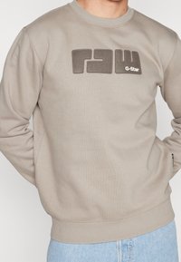 Felpa beige chiaro con logo "RAW" testurizzato in grigio scuro, scollatura rotonda, polsini e orlo a coste, e morbida texture del tessuto.