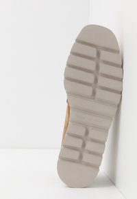Semelle d'une chaussure beige clair avec des rainures rectangulaires profondes, positionnée verticalement contre un fond d'angle blanc.